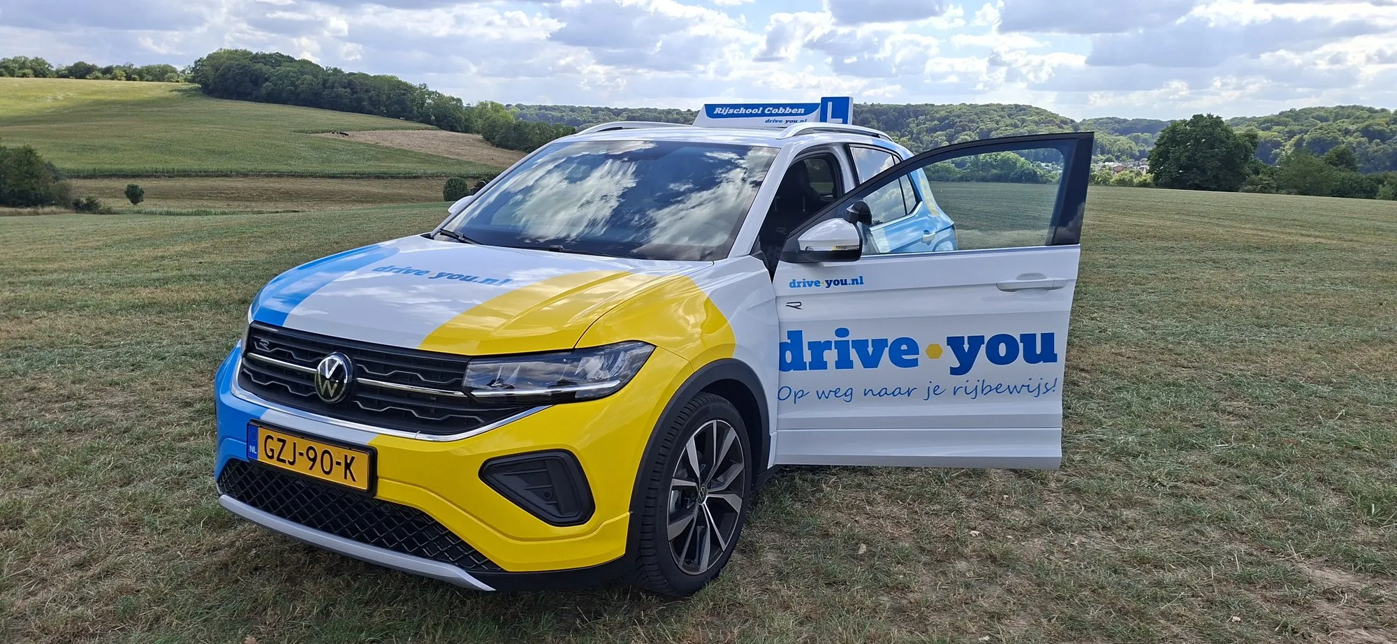 De lesauto van Rijschool Cobben, een witte Volkswagen T-Cross met drive.you-belettering, in het Zuid-Limburgse heuvellandschap