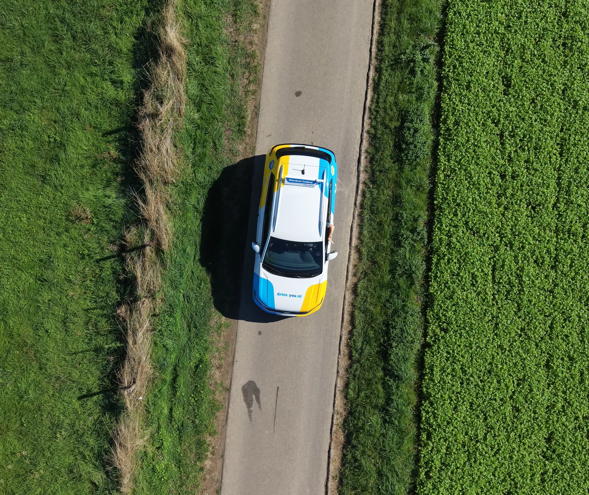 De lesauto van Rijschool Cobben van bovenaf gezien op een smalle landweg tussen weilanden in Zuid-Limburg, met Niels die zijn arm uit het raam steekt